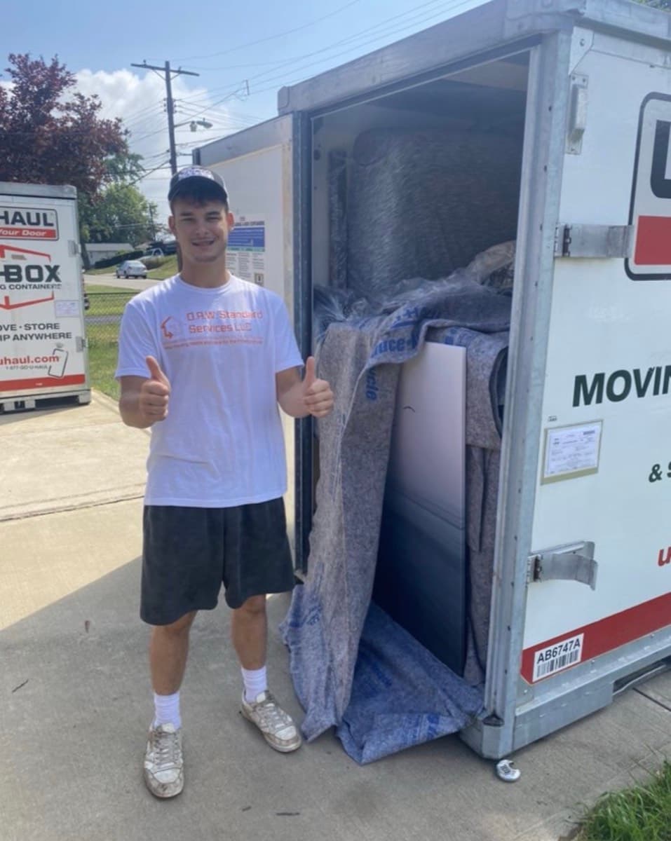 A DAW mover in a company shirt beside a moving container packed with blanket-wrapped furniture.